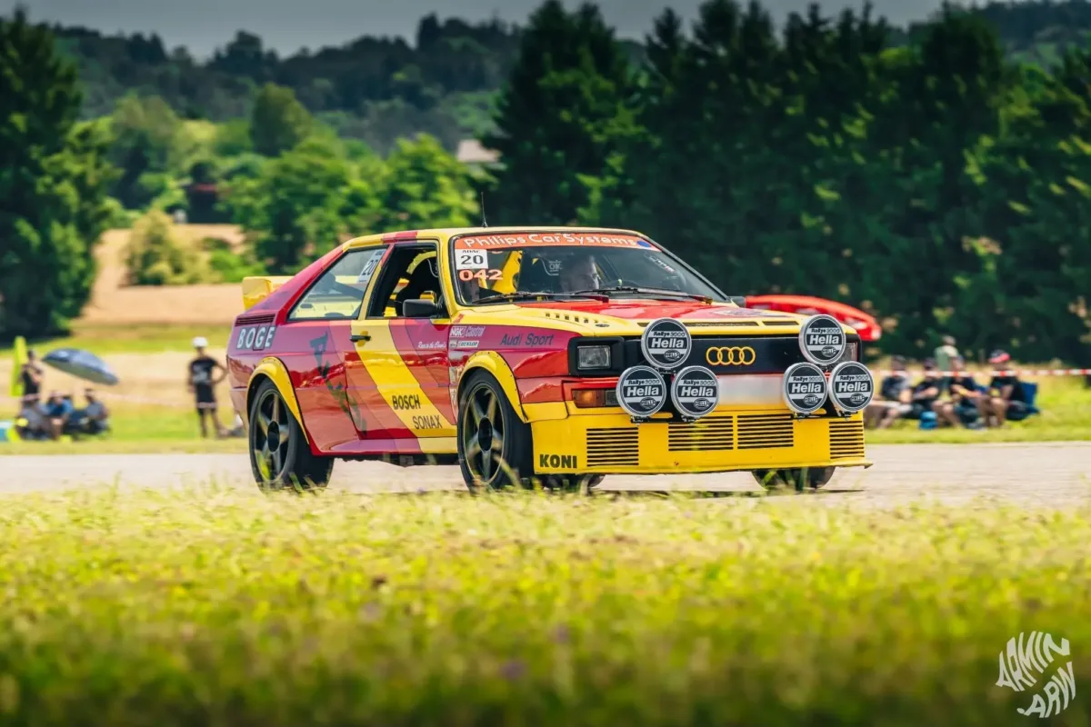 Rot-gelber Audi quattro mit Hella-Zusatzscheinwerfern in Fahrt bei einer Motorsportveranstaltung, im Hintergrund Zuschauer
