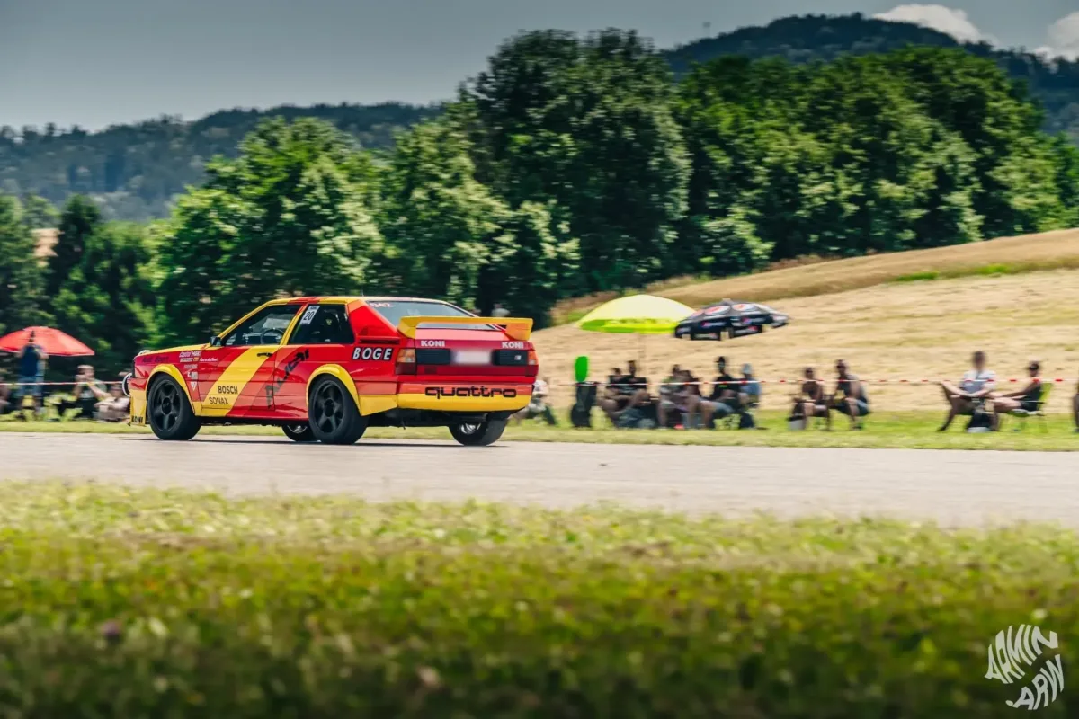 Audi quattro im Rallye-Design schräg von hinten in Fahrt, am Streckenrand sitzen Zuschauer