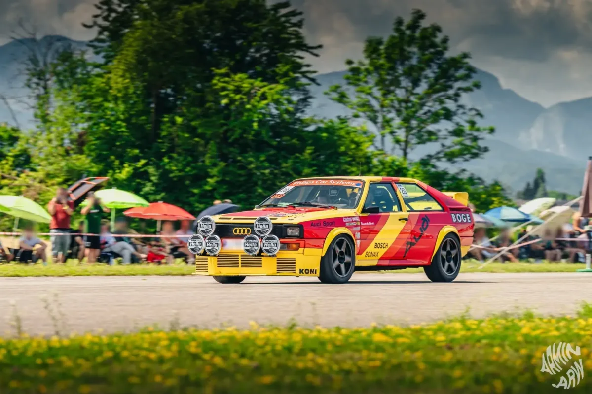 Audi quattro im rot-gelben Rallye-Design in Fahrt, dahinter Zuschauer mit Sonnenschirmen und Berge