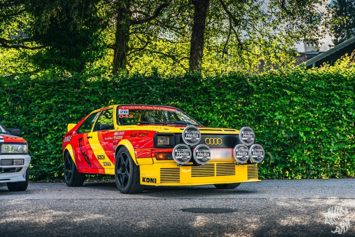 Audi quattro im rot-gelben Rallye-Design mit Hella-Zusatzscheinwerfern und KONI-Schriftzug, schräg von vorne vor einer Hecke
