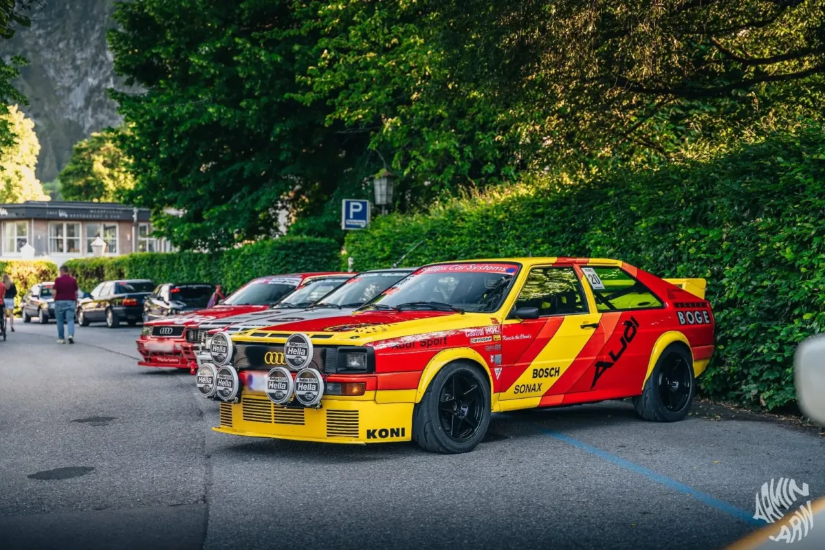 Audi quattro im rot-gelben Rallye-Design mit Hella-Zusatzscheinwerfern auf einem Parkplatz, dahinter weitere Audi quattro