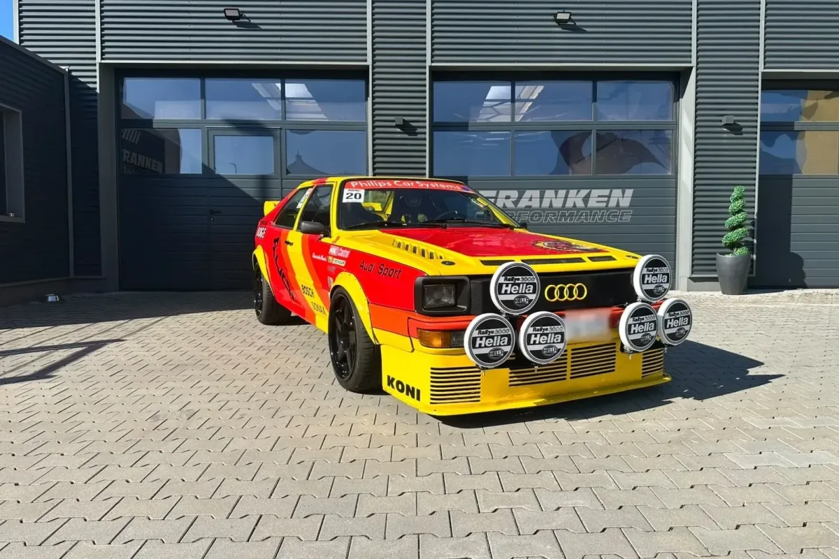 Audi quattro im rot-gelben Rallye-Design mit sechs Hella-Zusatzscheinwerfern und Startnummer 20 vor dem Werkstattgebäude von Franken Performance