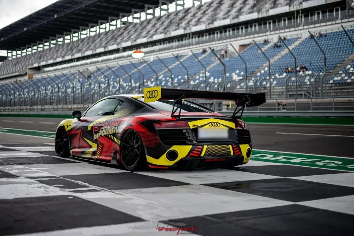 Audi R8 V10 Plus mit großem Heckflügel und Folierung im Franken-Performance-Design, schräg von hinten auf schwarz-weiß kariertem Boden vor der Tribüne einer Rennstrecke