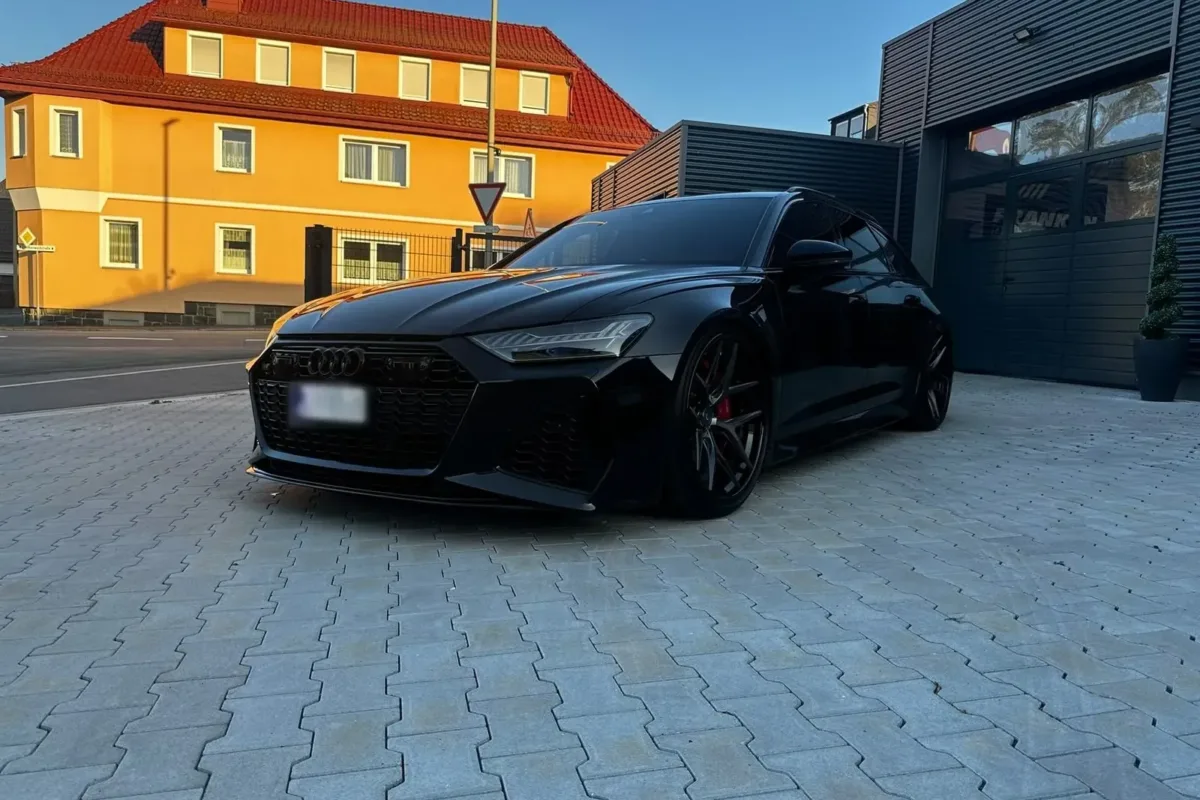 Schwarzer Audi RS6 C8 Avant schräg von vorne im Abendlicht vor dem Werkstattgebäude, im Hintergrund ein gelbes Wohnhaus mit rotem Dach