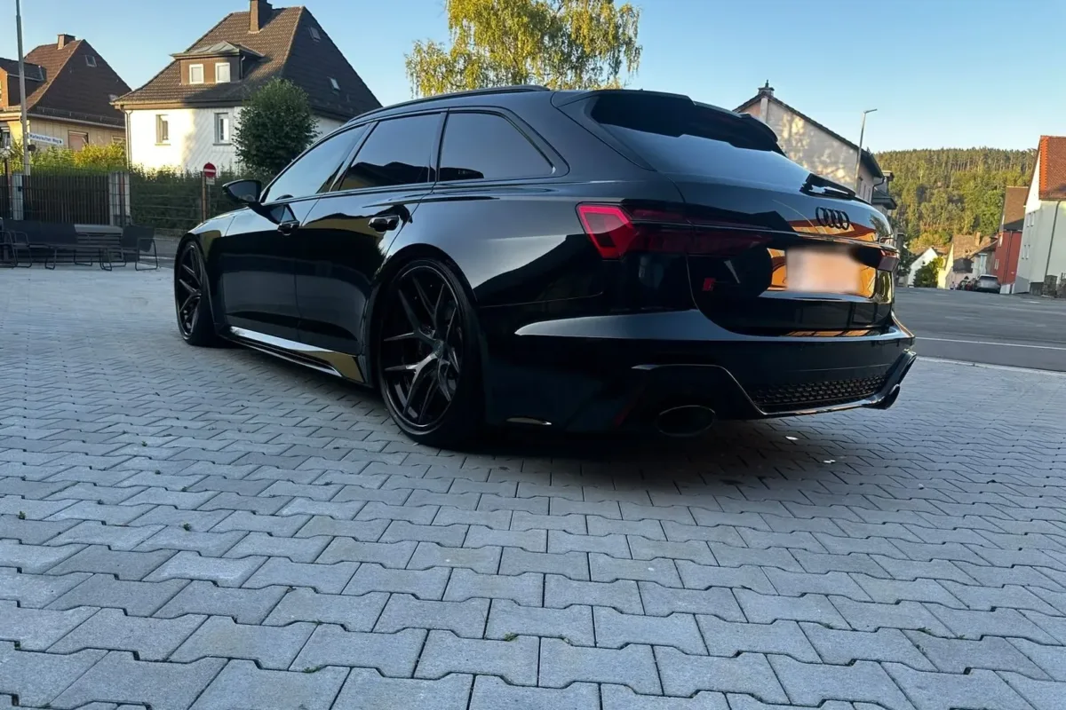 Heck des schwarzen Audi RS6 C8 Avant mit ovalen Endrohren und schwarzen Vossen-Felgen, schräg von hinten links auf dem Hof der Werkstatt
