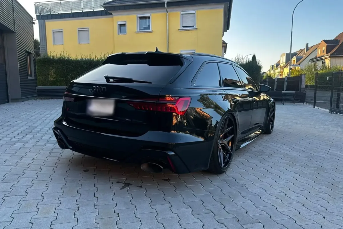 Schwarzer Audi RS6 C8 Avant schräg von hinten rechts mit durchgehendem LED-Leuchtenband und schwarzen Vossen-Felgen vor dem Werkstattgebäude