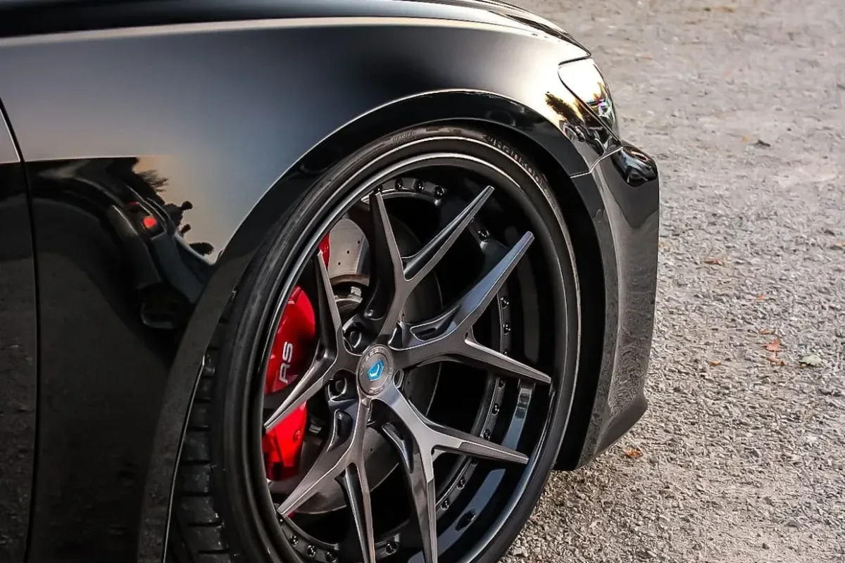 Detail des Audi RS6 C8: dreiteilige 21-Zoll-Vossen-Felge vor rotem Bremssattel, im Hintergrund der BMW E36 im Castrol-Design auf einem Tuning-Treffen