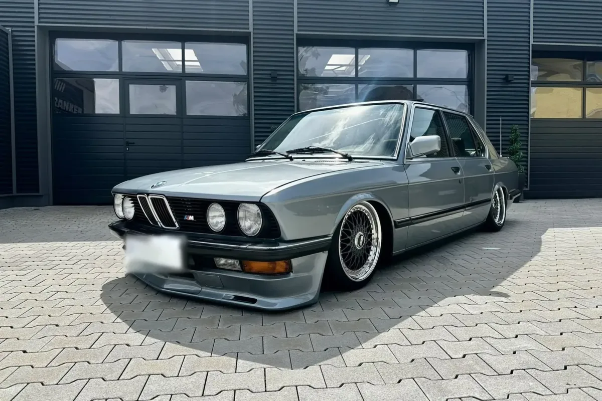 BMW E28 518i schräg von vorne links mit Doppelscheinwerfern, Niere mit M-Emblem und BBS-Felgen, auf Luftfahrwerk abgesenkt vor dem Werkstattgebäude
