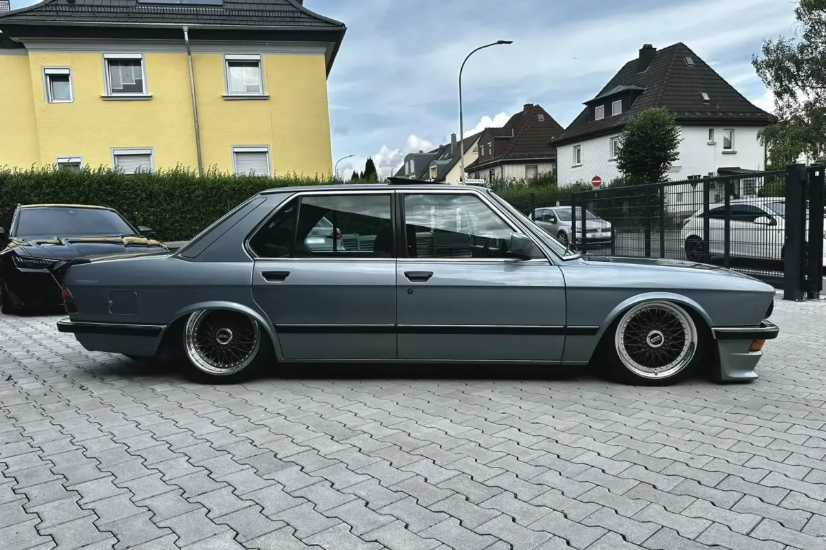 Seitenansicht des graublauen BMW E28 518i, auf Luftfahrwerk abgesenkt, mit BBS-Kreuzspeichenfelgen auf dem Hof der Werkstatt