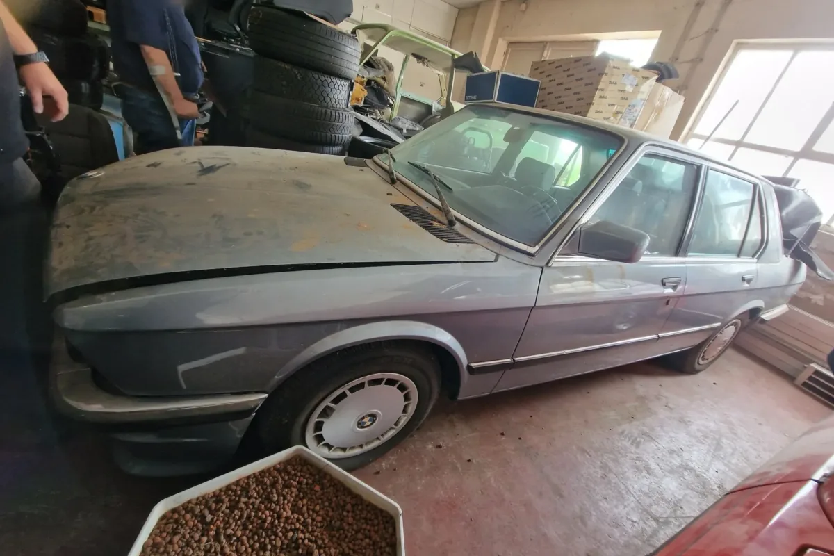 Der BMW E28 518i vor der Teilrestaurierung: verstaubt und mit originalen BMW-Radkappen in einer vollgestellten Halle zwischen Reifenstapeln und Kartons