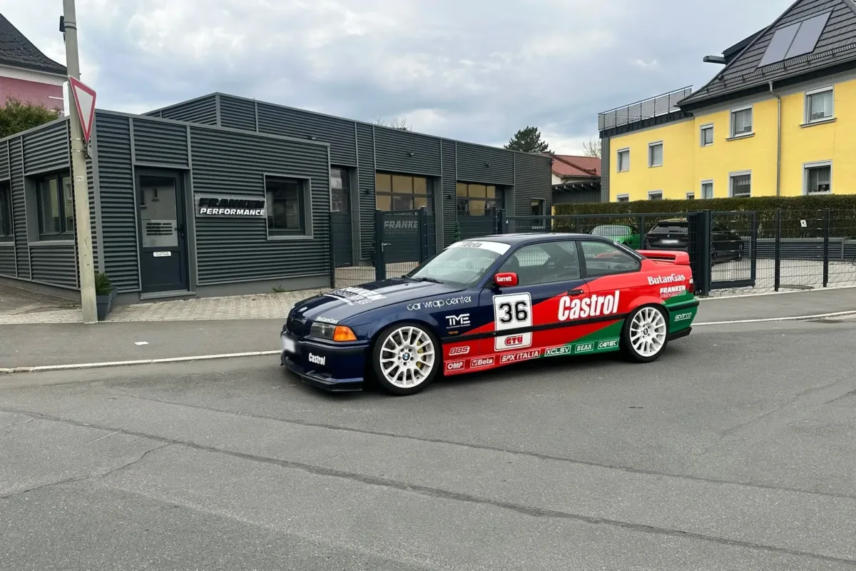 BMW E36 Coupé im Castrol-Design in Blau, Rot und Grün mit Startnummer 36 und weißen Felgen vor dem Werkstattgebäude von Franken Performance