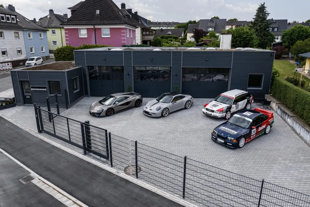 Luftaufnahme der Werkstatt von Franken Performance mit dunkler Metallfassade; auf dem Hof stehen ein McLaren, ein Porsche 911 (992) GT3 Touring und die Projektfahrzeuge Audi S6 C4 Avant und BMW E36 Coupé im Motorsport-Design