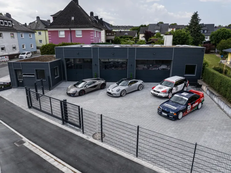 Luftaufnahme der Werkstatt von Franken Performance mit dunkler Metallfassade; auf dem Hof stehen ein McLaren, ein Porsche 911 (992) GT3 Touring und die Projektfahrzeuge Audi S6 C4 Avant und BMW E36 Coupé im Motorsport-Design