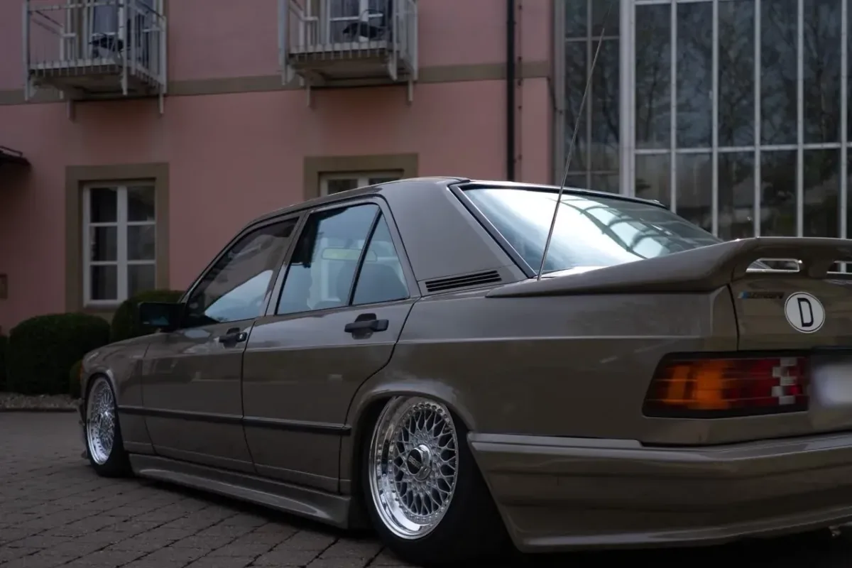 Heck des Mercedes-Benz 190 (W201) mit Heckspoiler und BBS-RS-Felgen, schräg von hinten vor einem rosafarbenen Gebäude mit Glasfassade