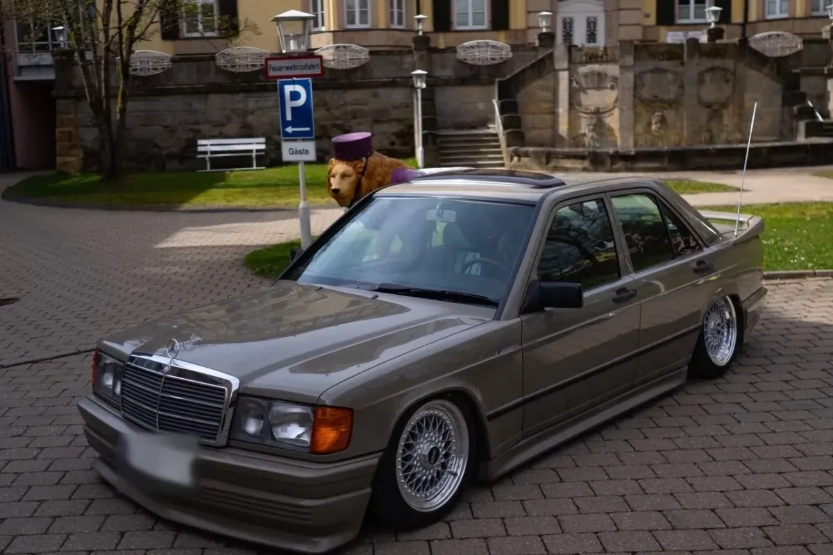 Mercedes-Benz 190 (W201) mit Zender-Frontschürze, auf Luftfahrwerk abgesenkt, schräg von vorne auf einem gepflasterten Platz vor einem historischen Gebäude