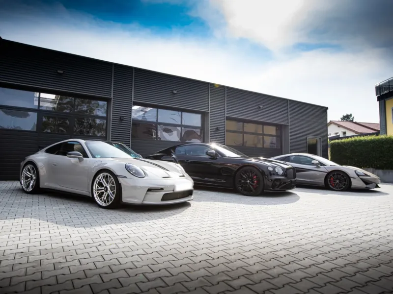 Grauer Porsche 911 (992) GT3 Touring, schwarzer Bentley Continental GT und silberner McLaren vor den Hallentoren des dunklen Werkstattgebäudes von Franken Performance